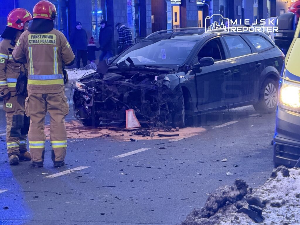 Strażacy w mundurach z logo PSP oceniają uszkodzenia samochodu po wypadku na miejskiej ulicy, obok leżą resztki pojazdu.