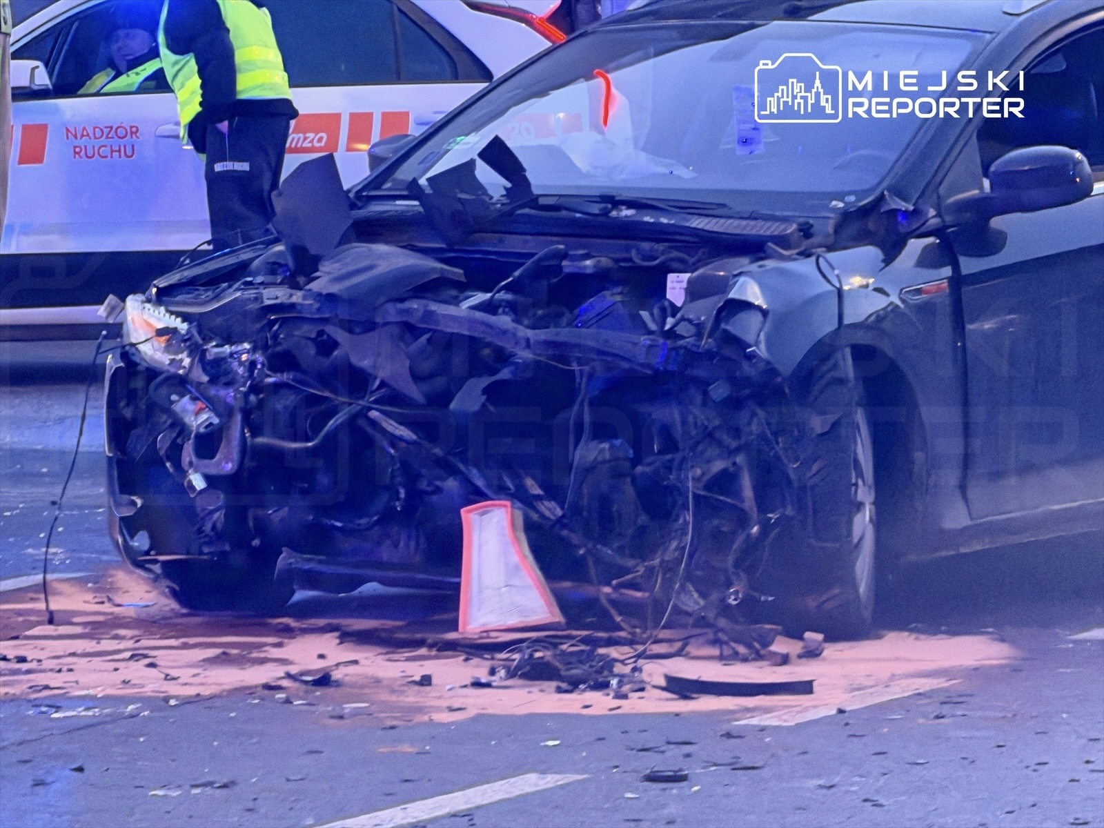 Zniszczony samochód osobowy z uszkodzoną przednią częścią leży na jezdni, obok stoi policjant w kamizelce odblaskowej.