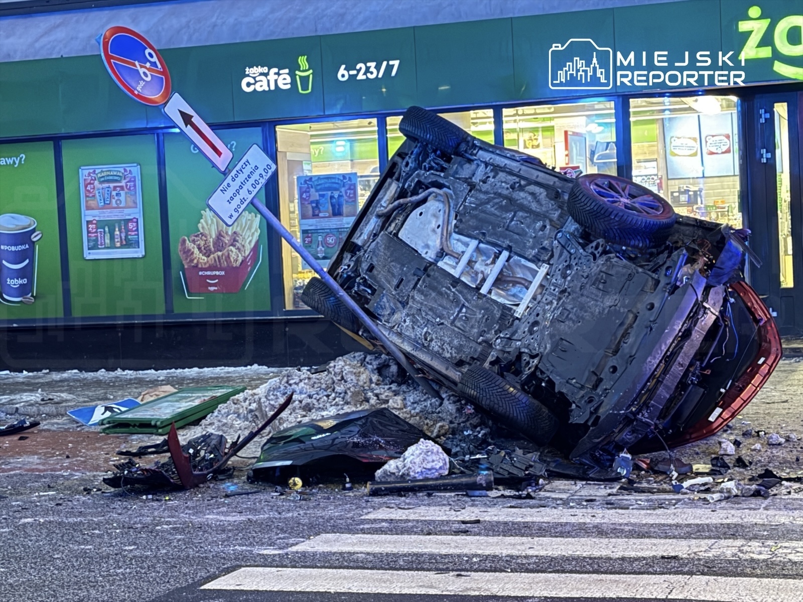 Przewrócone auto leży na boku obok znaku zakazu parkowania, w tle widoczny sklep spożywczy.