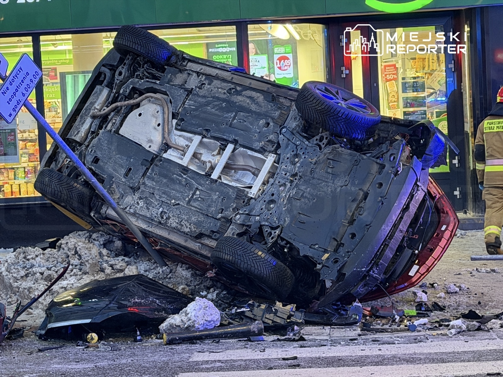 Zniszczony samochód leży na dachu obok znaku drogowym, w tle widoczny sklep oraz strażacy w mundurach.