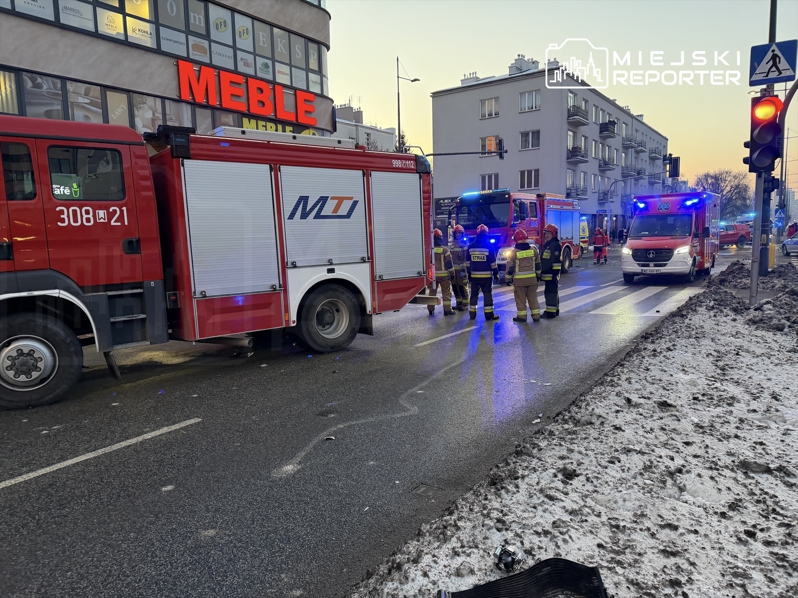 Strażacy w mundurach omawiają sytuację przy czerwonym wozie strażackim obok oświetlonego budynku z napisem "MEBLE".