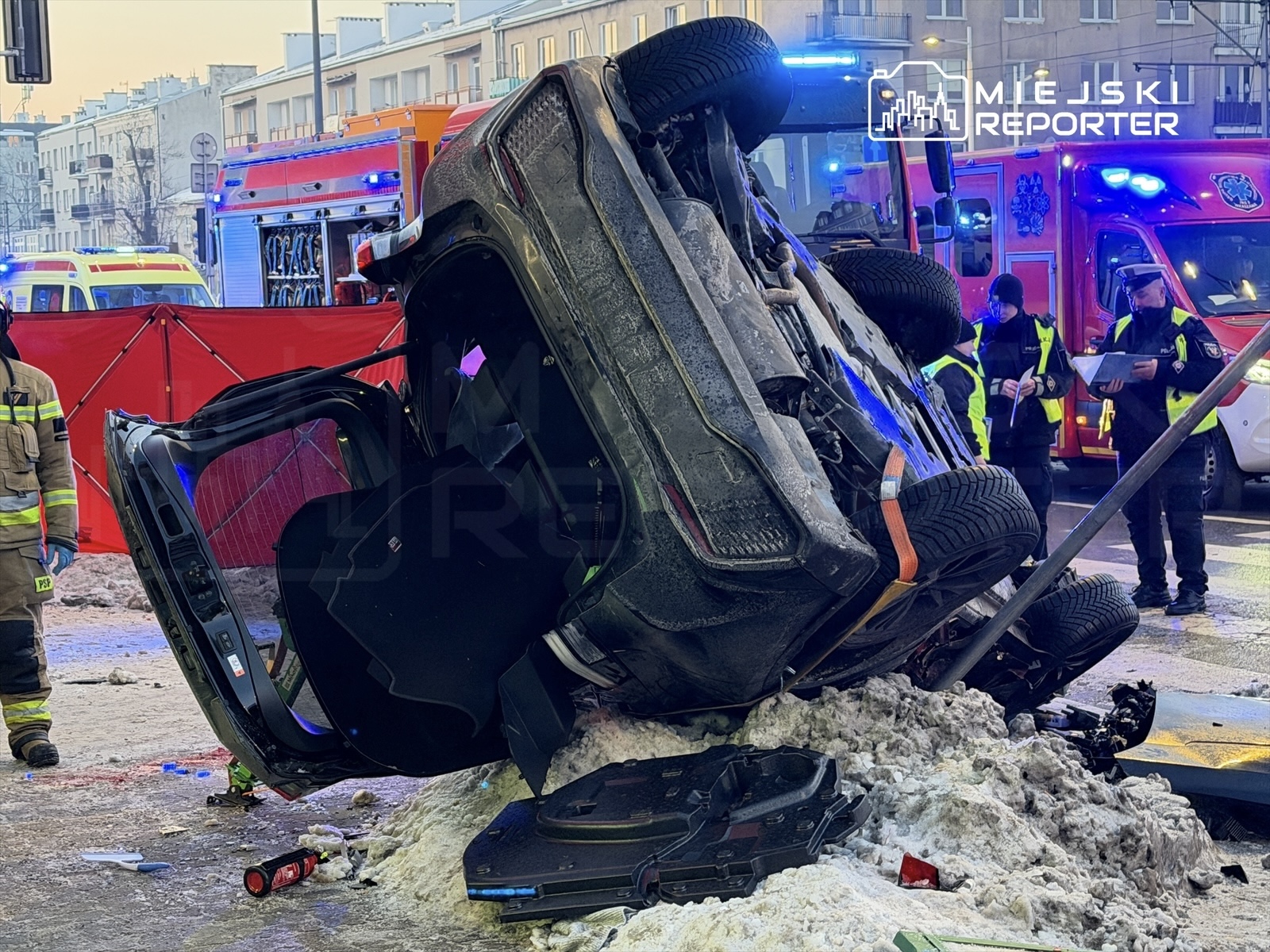 Samochód leżący na dachu na zaśnieżonej ulicy, w tle strażacy i policjanci prowadzący działania ratunkowe.