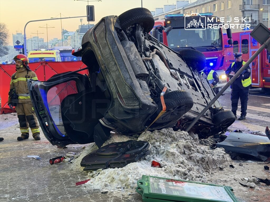 Samochód leżący na dachu na zaśnieżonej ulicy, obok strażak w sprzęcie ochronnym oraz policjant badający miejsce zdarzenia.