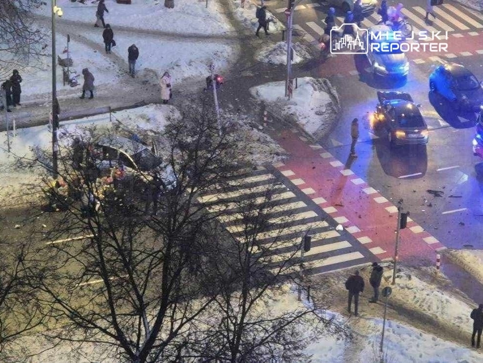 Na skrzyżowaniu ulic, kilka osób otacza uszkodzony samochód, a w tle widać ruch uliczny oraz śnieg.