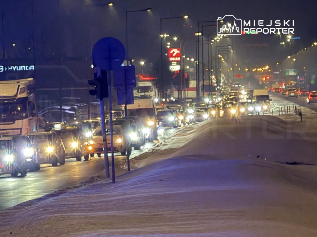 Ruch samochodowy na ośnieżonej ulicy w nocy, z widocznymi światłami pojazdów i lampami ulicznymi.