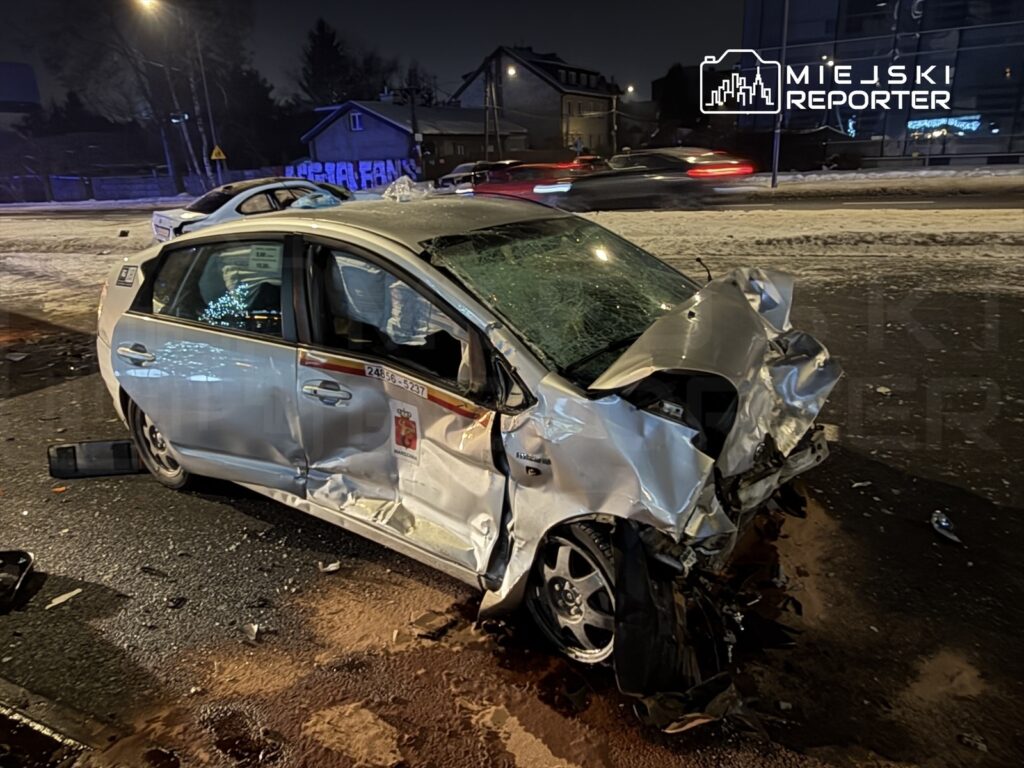 Rozbity taksówka z uszkodzoną przednią częścią stoi na ośnieżonej jezdni w nocy, otoczona przez inne pojazdy.