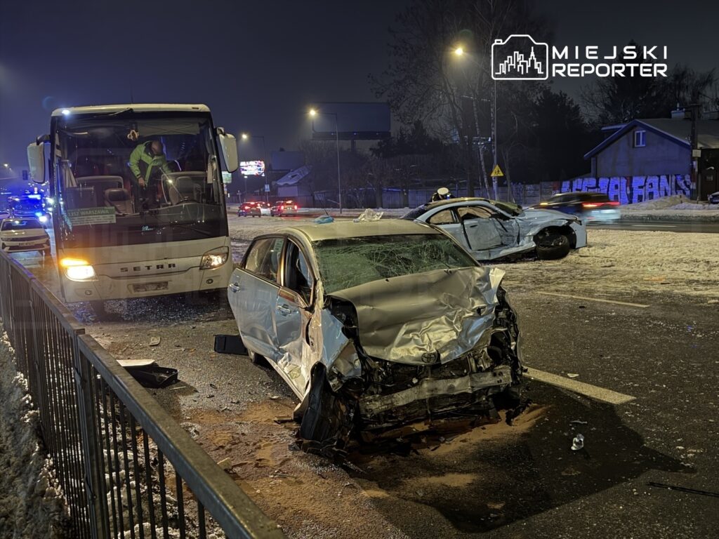 W nocy na drodze znajduje się uszkodzony samochód osobowy, a obok stoi autobus oraz rozbite auto.