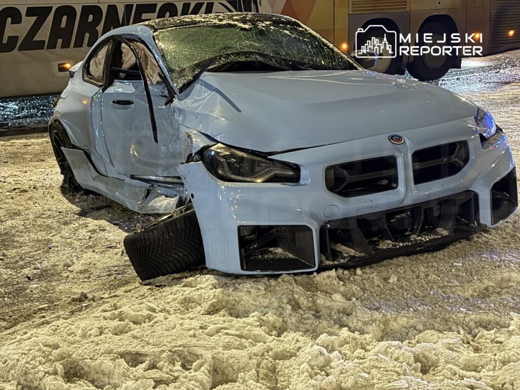 Uszkodzony samochód sportowy z wgniecioną karoserią stoi na zaśnieżonej nawierzchni, obok widoczny pojazd autobusowy.