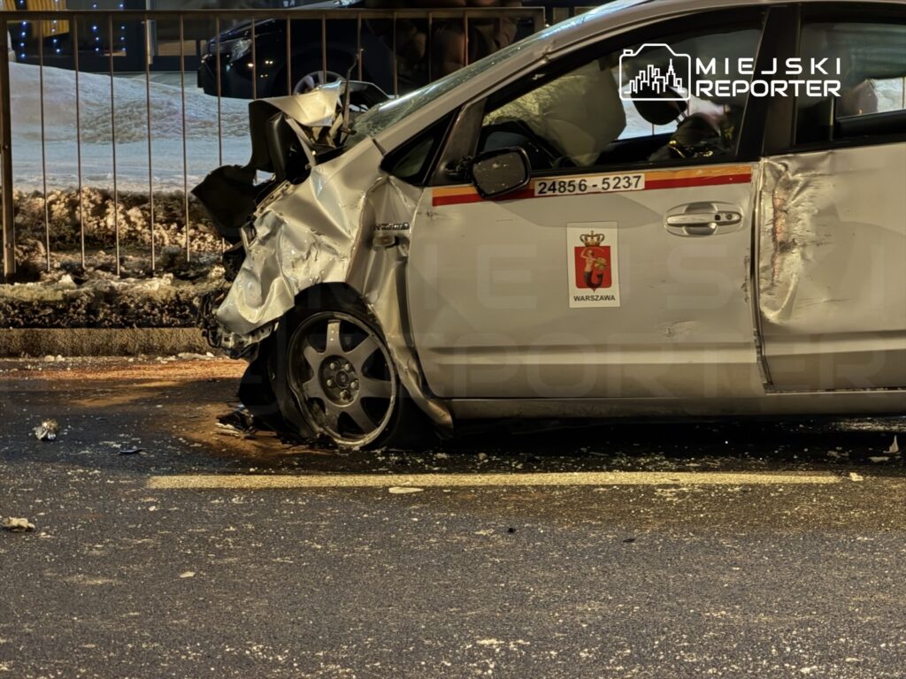 Uszkodzony samochód osobowy z widoczną wgniecioną tylną częścią stoi na ulicy, obok ogrodzenia.
