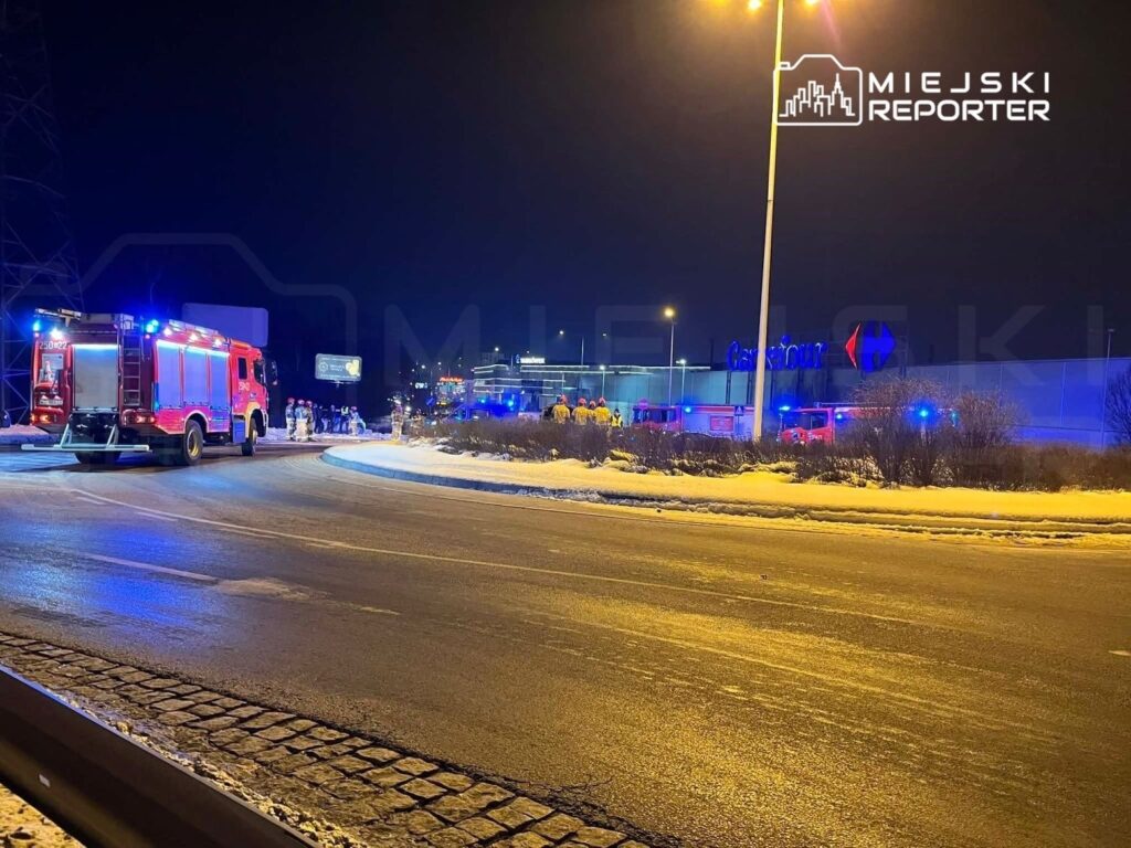 W nocy przy rondzie, w pobliżu supermarketu, strażacy interweniują w akcji ratunkowej, używając wozów strażackich.