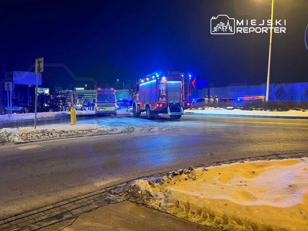 Na skrzyżowaniu w nocy stoją wozy strażackie i ambulans, a w tle widać grupę ratowników.