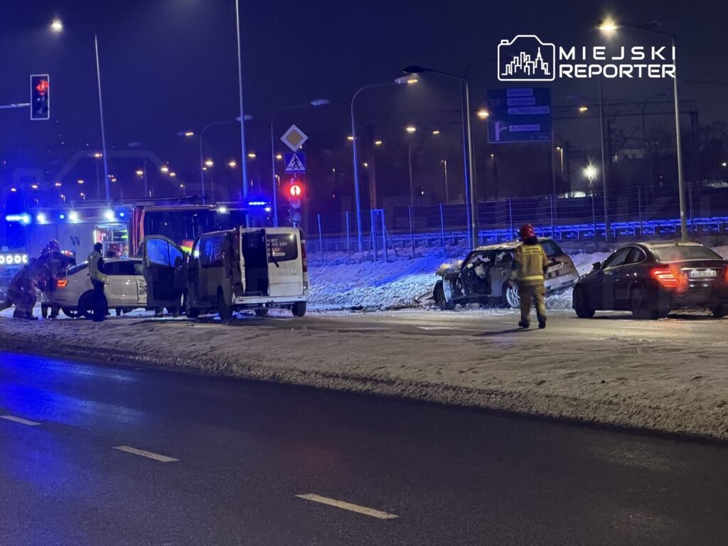 Ratownicy w mundurach sprawdzają uszkodzone samochody na zaśnieżonej drodze w nocy, w tle widać sygnały świetlne.