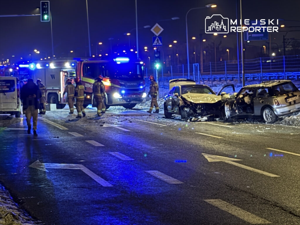 Strażacy pracują przy wypadku drogowym, obok uszkodzonych samochodów na oświetlonej ulicy nocą.