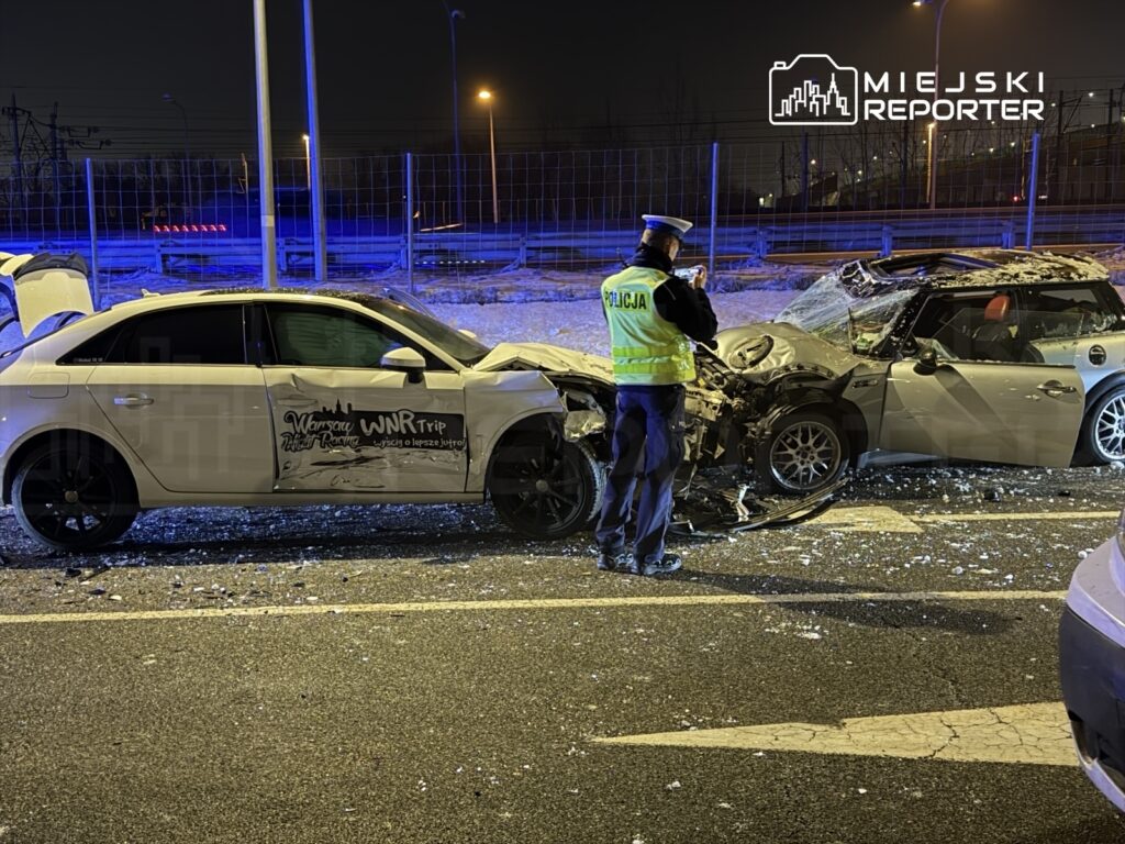Policjant dokumentuje wypadek drogowy z udziałem dwóch uszkodzonych samochodów na nocnej jezdni.