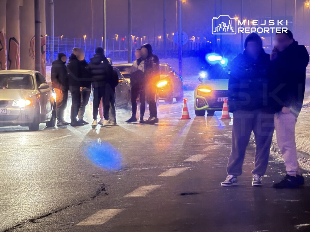 Grupa mężczyzn rozmawia na ulicy obok zaparkowanych samochodów, w tle świecą niebieskie światła radiowozu.