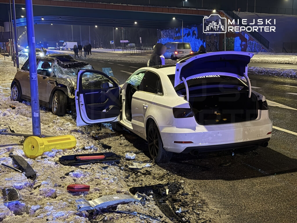 Dwa uszkodzone samochody stoją na poboczu drogi, wokół leżą fragmenty wraku i żółty pojemnik.