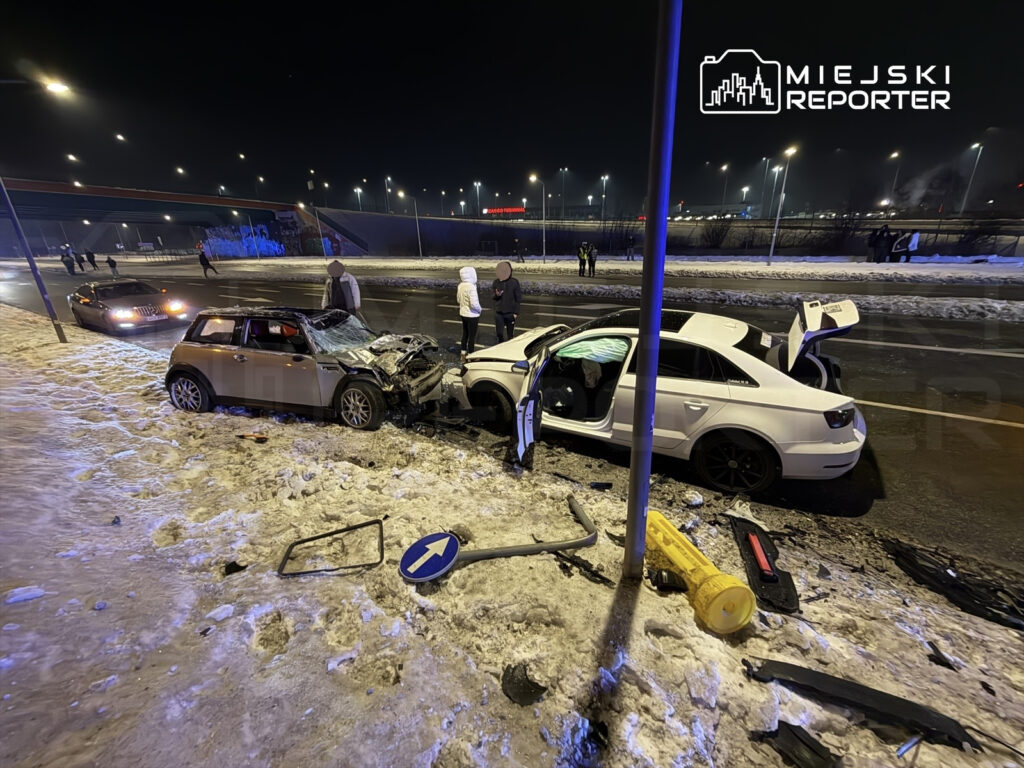 Dwa uszkodzone pojazdy na drodze, wokół zbierają się ludzie, a w tle widać oświetlone miasto.