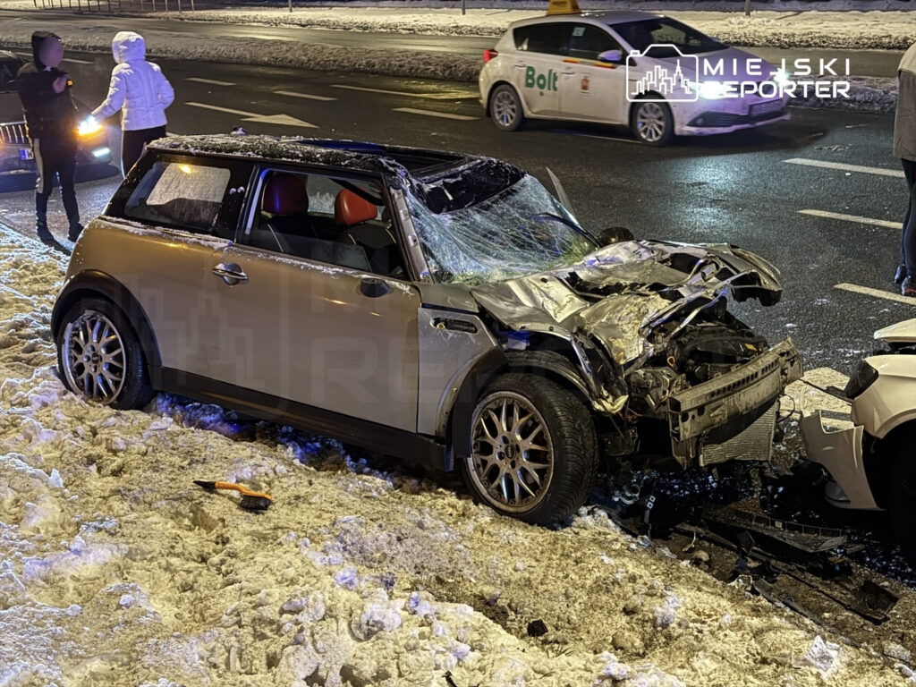Zniszczony samochód osobowy zaparkowany na ośnieżonym poboczu, obok dwie osoby rozmawiające w nocy.