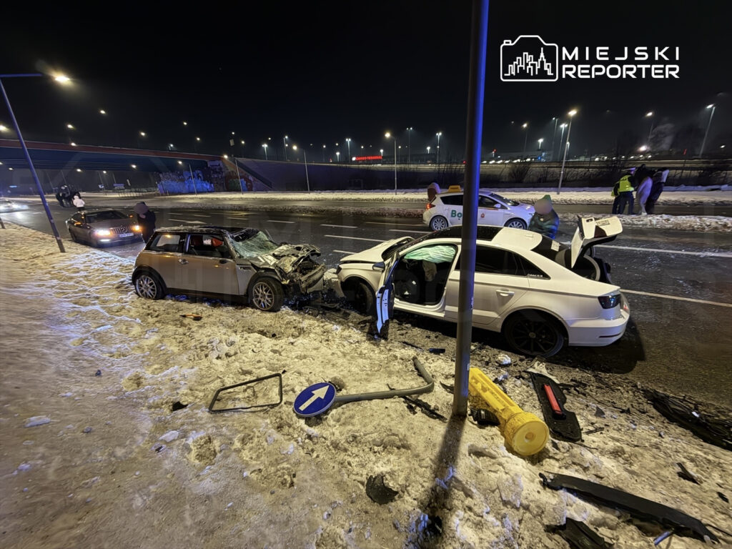 Dwa uszkodzone samochody po zderzeniu na ośnieżonej jezdni, obok stoją policjanci oraz przechodnie.