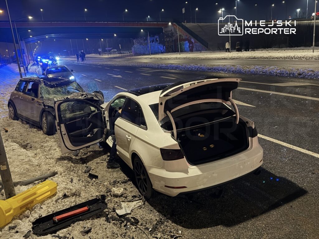 Dwa uszkodzone samochody na drodze, jeden z otwartymi drzwiami, w nocy, w tle widać niebieskie światła radiowozów.