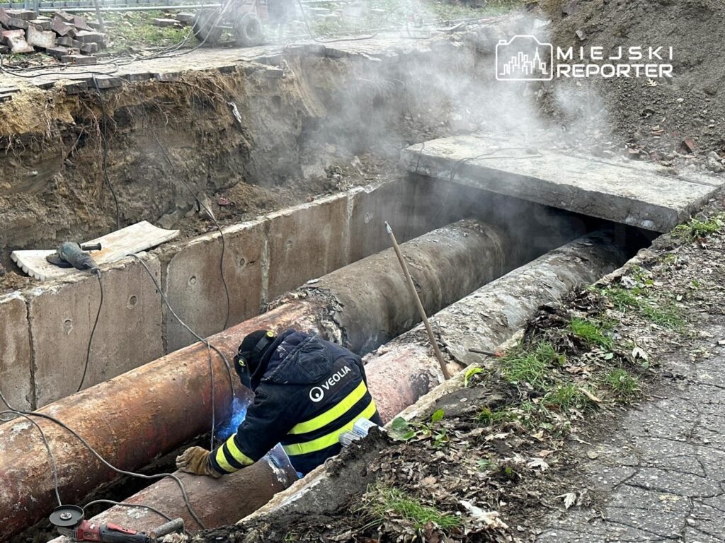 Technik w odzieży roboczej spawa rury w wykopie, emitując niebieski łuk elektryczny, otoczony przez wydobywający się dym.