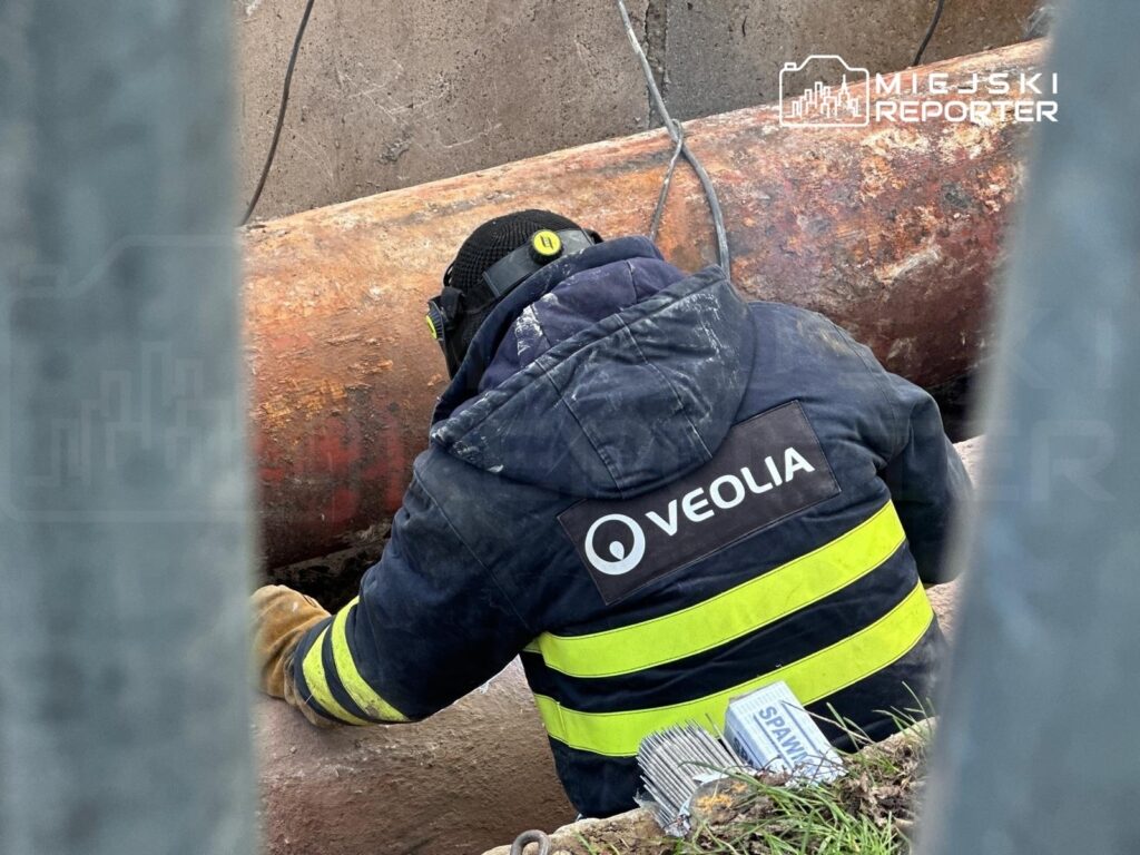 Pracownik w odzieży roboczej firmy Veolia naprawia rury w wykopie, obok leżą materiały spawalnicze.