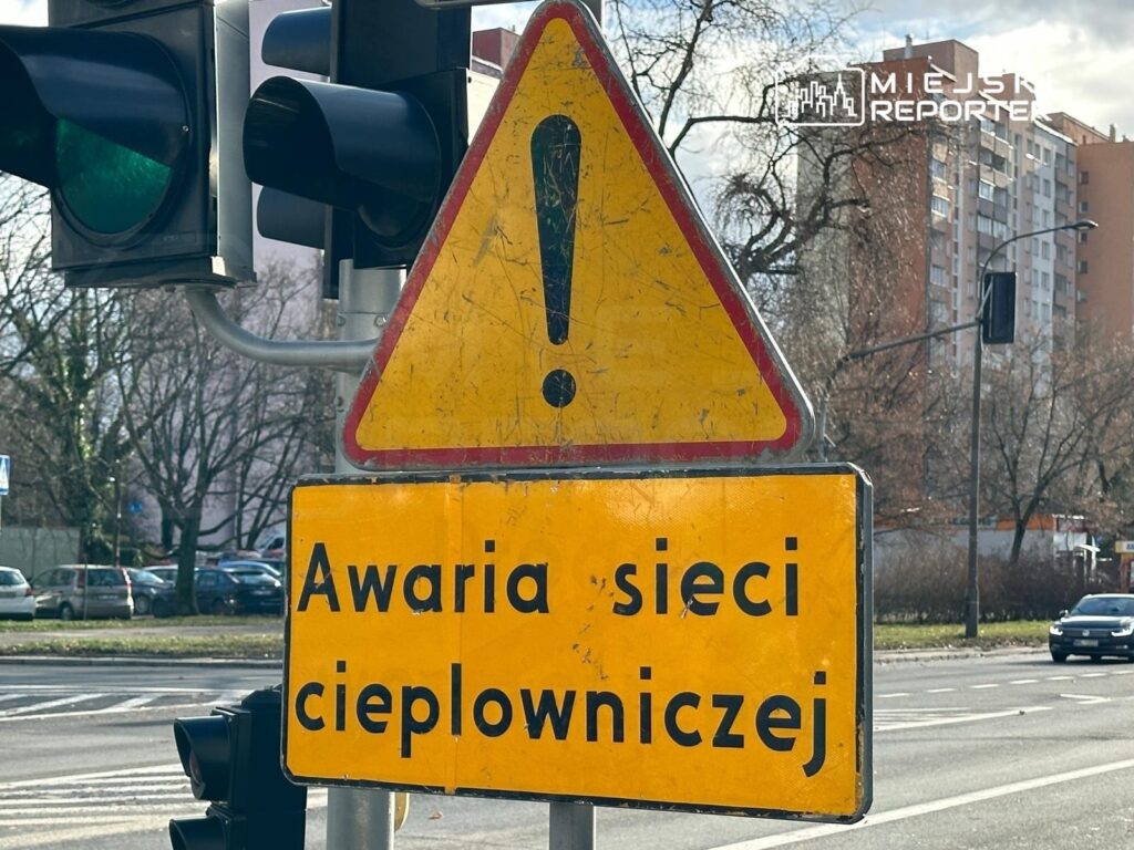 Żółty znak ostrzegawczy informujący o awarii sieci ciepłowniczej, umieszczony przy ulicy obok sygnalizacji świetlnej.