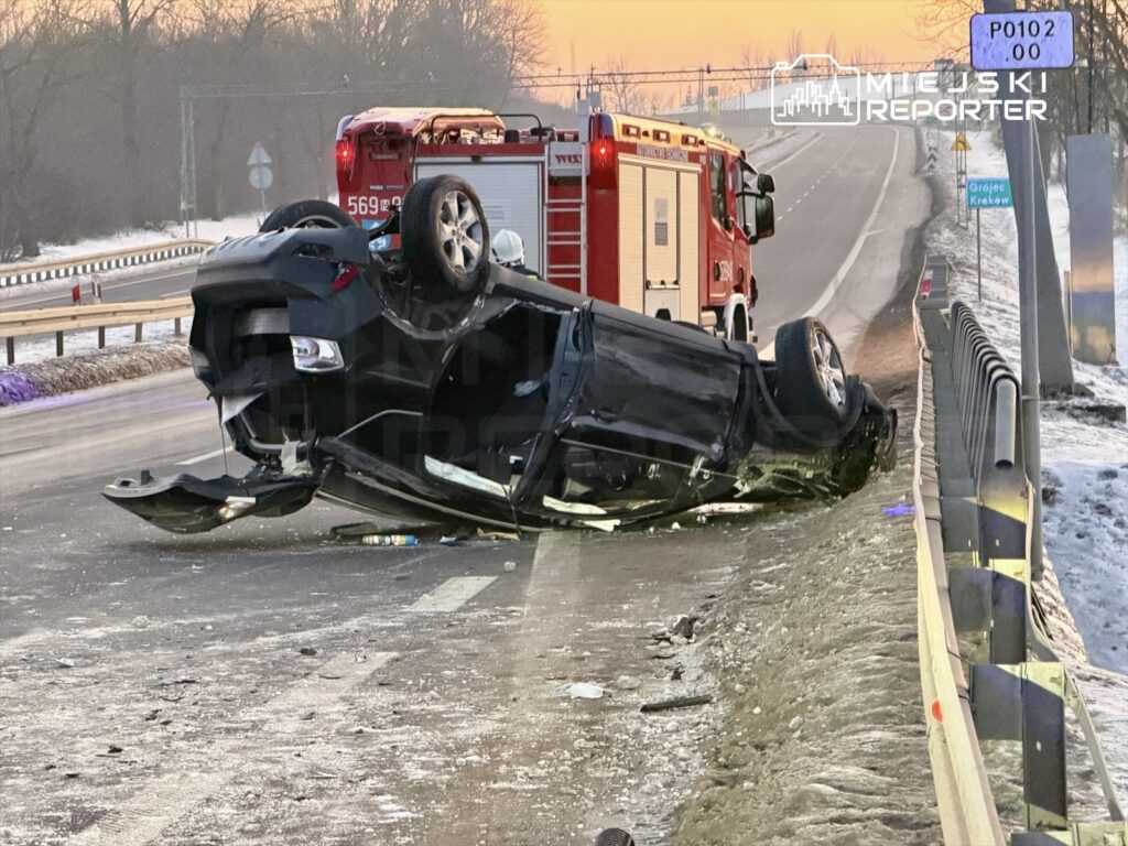 Czarny samochód osobowy leży na dachu przy drodze, obok stoi wóz strażacki, w tle widoczne drzewa i sygnalizacja drogowa.