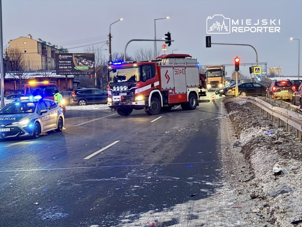 Wozidło straży pożarnej oraz radiowóz policji na zablokowanej drodze, obok wypadku samochodowego w zimowym otoczeniu.