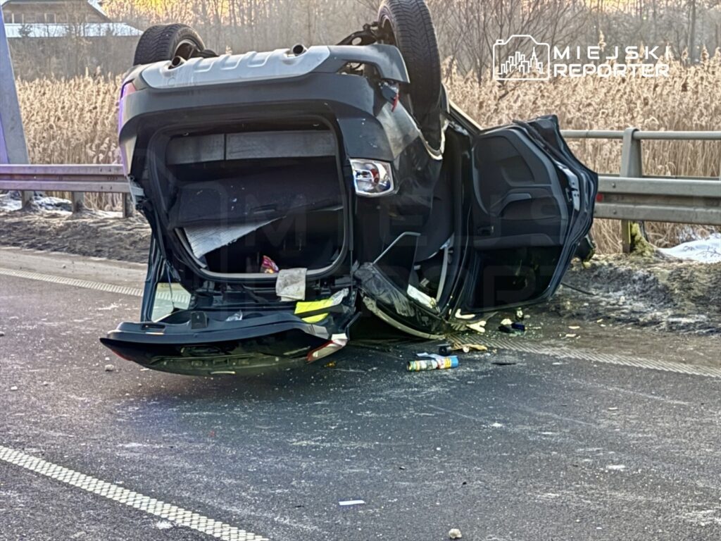 Samochód osobowy leży na dachu na poboczu drogi, otoczony rozrzuconymi przedmiotami.