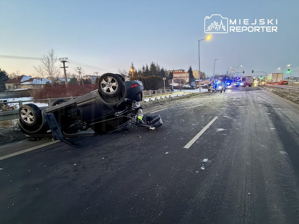 Przewrócone auto osobowe leży na drodze, obok widać pojazdy służb ratunkowych oraz zaspę śnieżną.