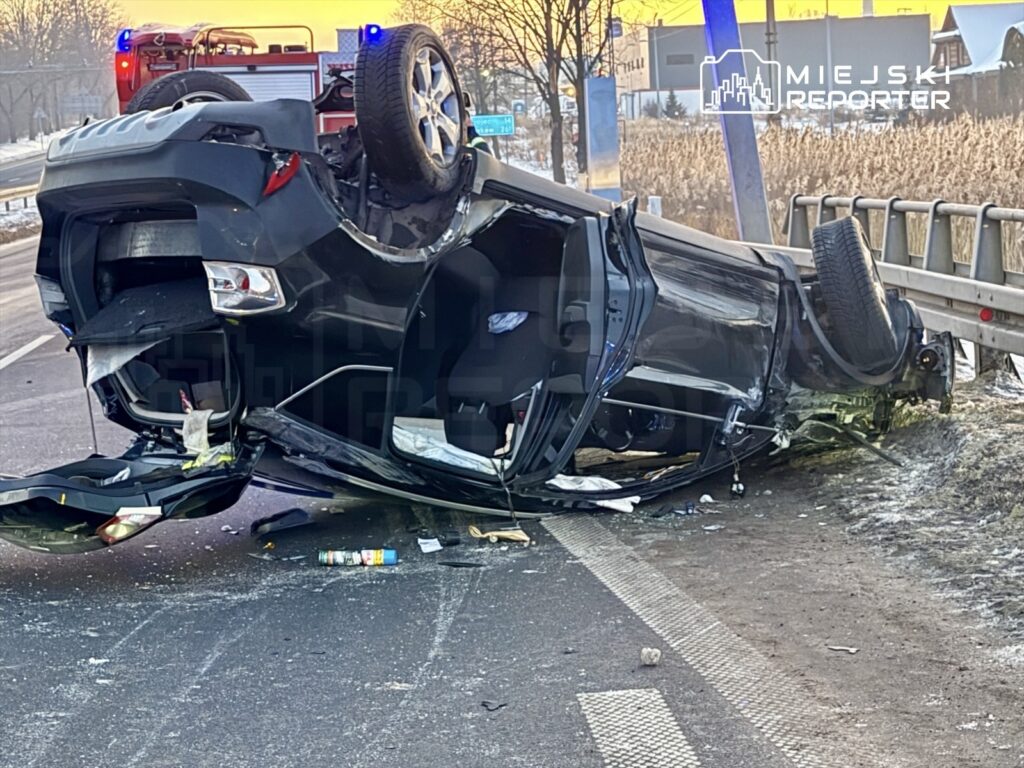 Czarny samochód osobowy leży na dachu obok drogi, z otwartymi drzwiami i widocznymi uszkodzeniami.