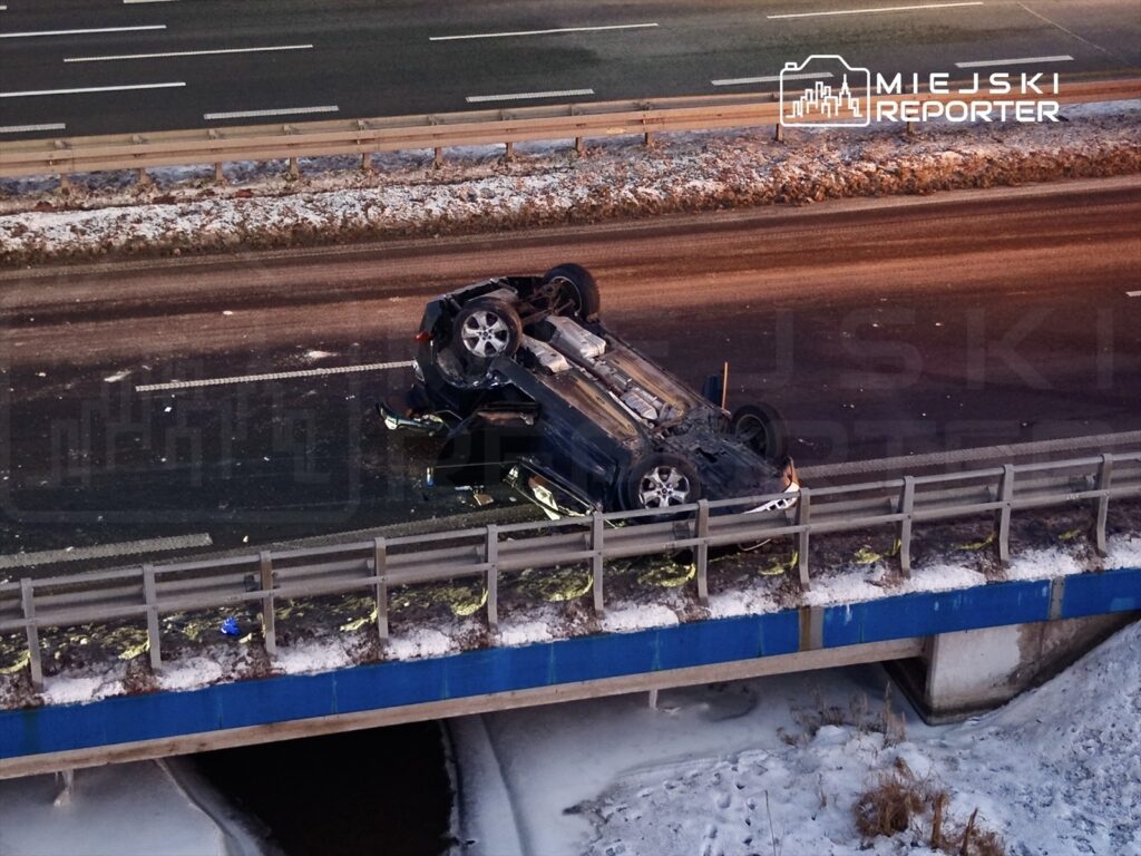 Samochód leżący na dachu na pasie drogowym, obok zamarzniętego pobocza, w tle widoczny ruch uliczny.