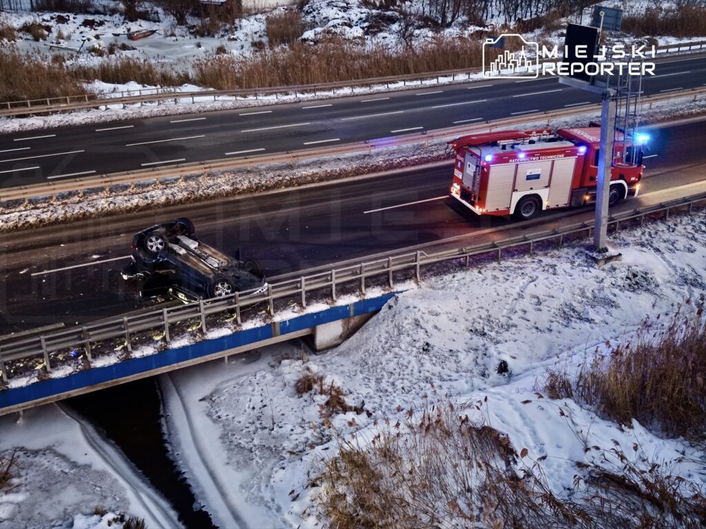 Na drodze leży przewrócone auto, a obok niego stoi wóz strażacki z włączonymi sygnałami świetlnymi.