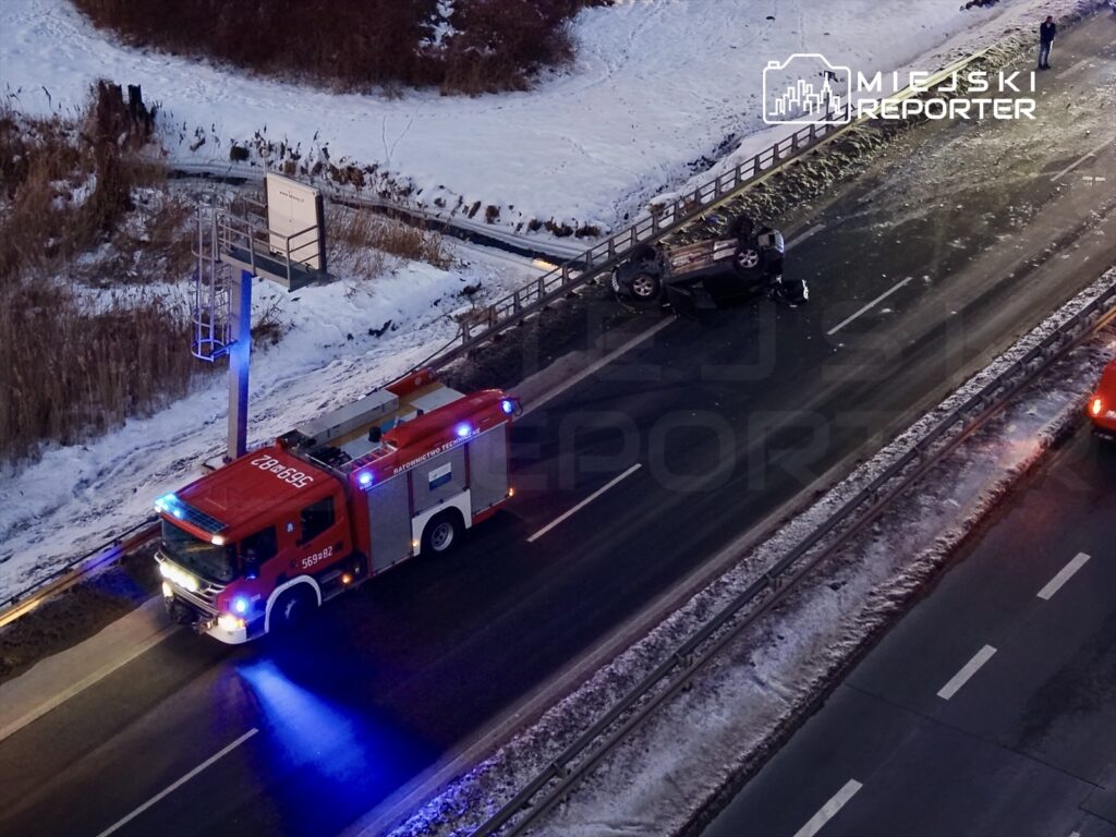 Czerwony wóz strażacki z włączonymi sygnalizatorami przy przewróconym samochodzie na drodze w zimowej scenerii.