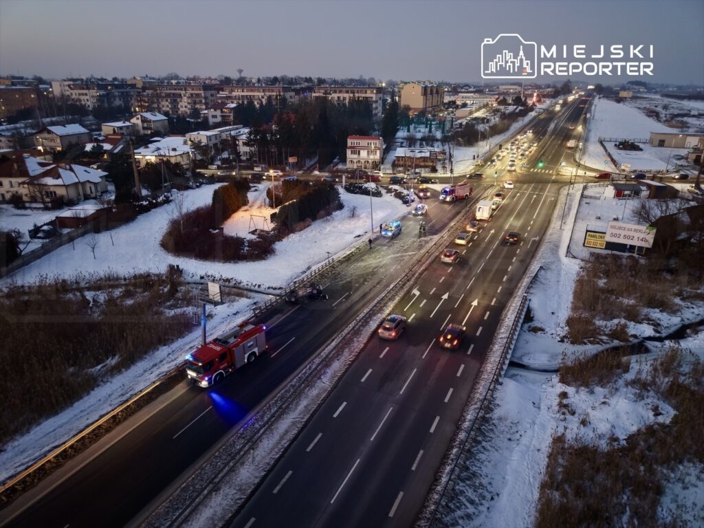 Na drodze w zimowym otoczeniu porusza się wóz strażacki z włączonymi sygnałami świetlnymi, obok przejeżdżają inne pojazdy.