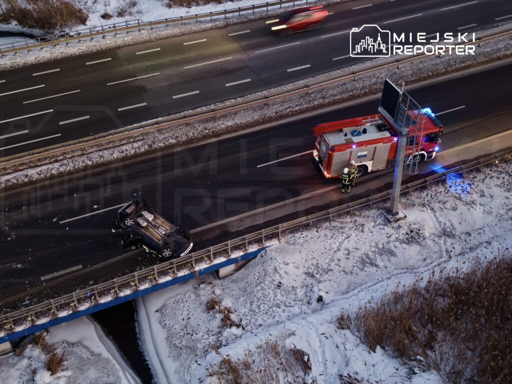 Na drodze leży przewrócone auto, obok stoi wóz strażacki z włączonymi sygnałami świetlnymi, a strażacy oceniają sytuację.