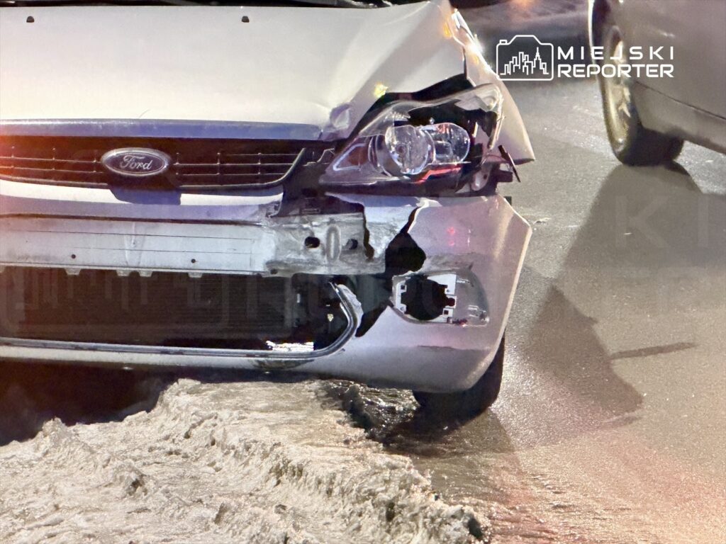 Uszkodzony biały samochód marki Ford z wgniecionym przodem stoi na ośnieżonej ulicy.