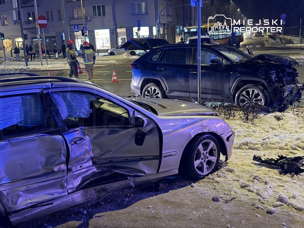 Dwa uszkodzone samochody osobowe na skrzyżowaniu, obok strażaków zabezpieczających miejsce zdarzenia.
