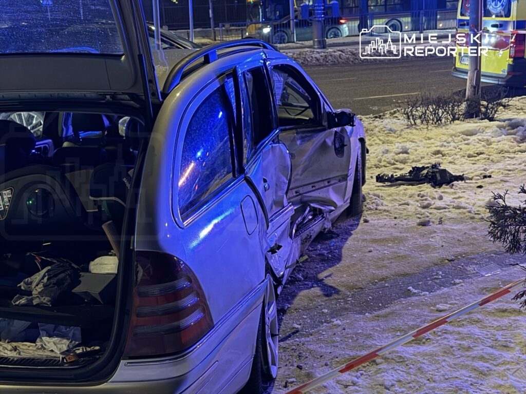 Uszkodzony samochód zaparkowany na chodniku, z otwartymi drzwiami, obok leży fragment pojazdu. W tle widać policyjne światła.