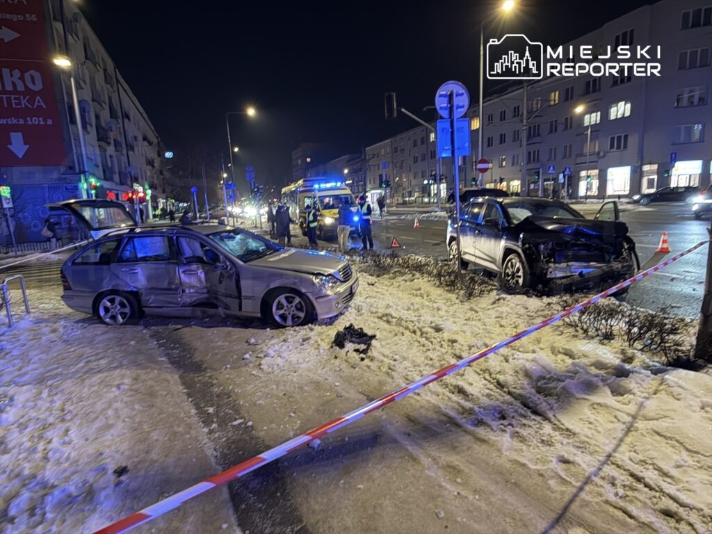 Dwa uszkodzone samochody osobowe na ulicy, w tle interweniujący ratownicy i radiowozy policyjne.