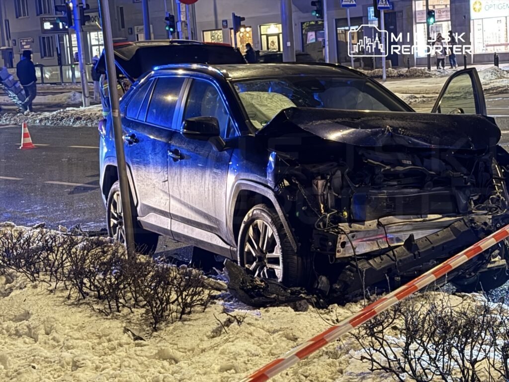 Uszkodzony samochód osobowy stoi przy ulicy, z otwartą klapą bagażnika, w otoczeniu śniegu i oznaczeń policyjnych.