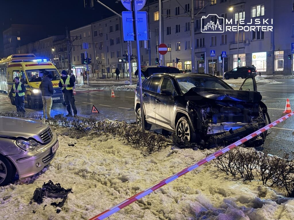 Na ulicy, w nocy, ratownicy medyczni oraz policjanci rozmawiają obok uszkodzonego samochodu osobowego.