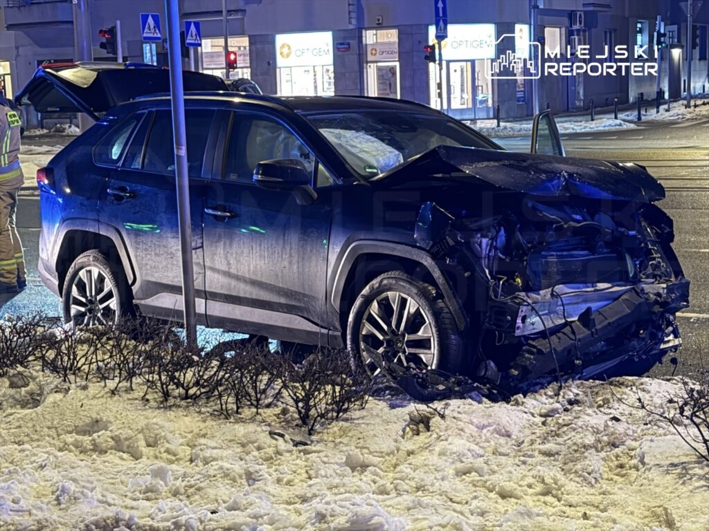 Czarny SUV z uszkodzoną tylną częścią stoi na poboczu drogi, obok strażaków badających sytuację.