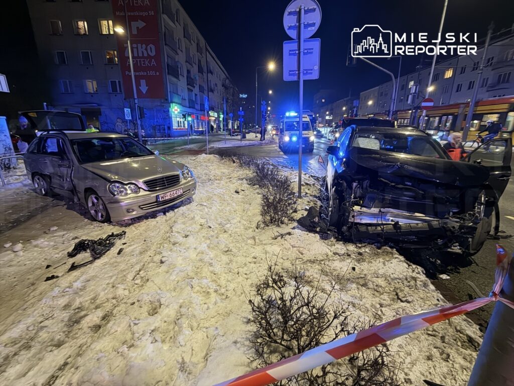 Dwa uszkodzone samochody osobowe na zaśnieżonej ulicy, w tle widać niebieskie światła radiowozu i budynki.