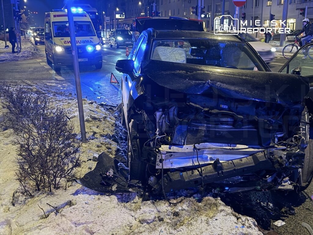 Rozbity samochód osobowy z uszkodzoną przednią częścią stoi na zaśnieżonym poboczu, obok widać ambulans i inne pojazdy.