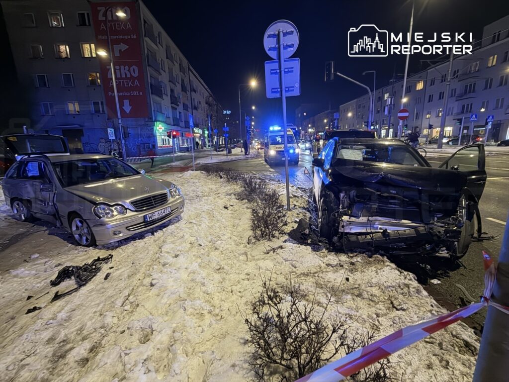 Dwa uszkodzone samochody osobowe stoją na chodniku, obok widoczne są pojazdy służb ratunkowych oraz oświetlenie uliczne.