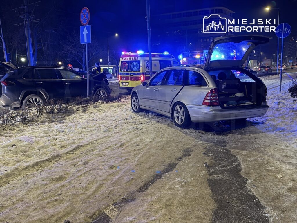 W nocy, na oświetlonej ulicy, dwa samochody po kolizji, jeden z otwartą klapą bagażnika, w tle karetka pogotowia.