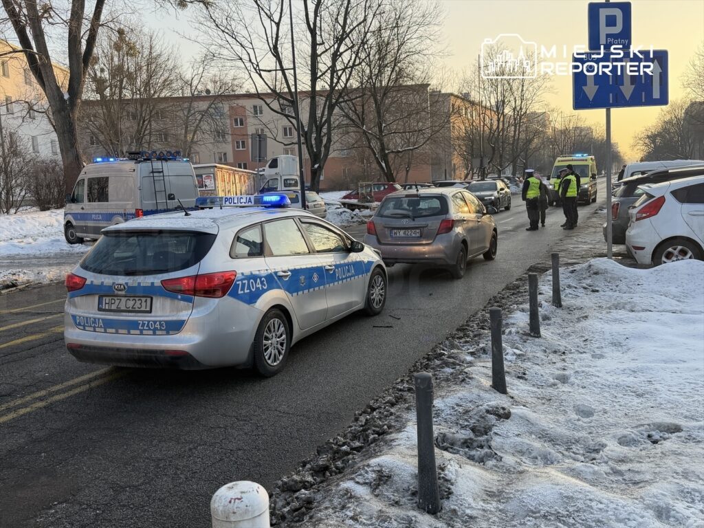 Policyjny radiowóz z włączonymi sygnałami świetlnymi stoi na ulicy, podczas gdy funkcjonariusze rozmawiają z kierowcą. W tle widać inne pojazdy oraz śnieżne pobocze.