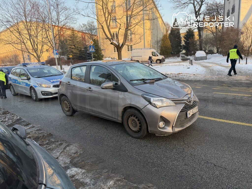 Na ulicy stoi szary samochód osobowy obok policyjnego radiowozu, a policjant rozmawia z kierowcą. W tle widoczne budynki.